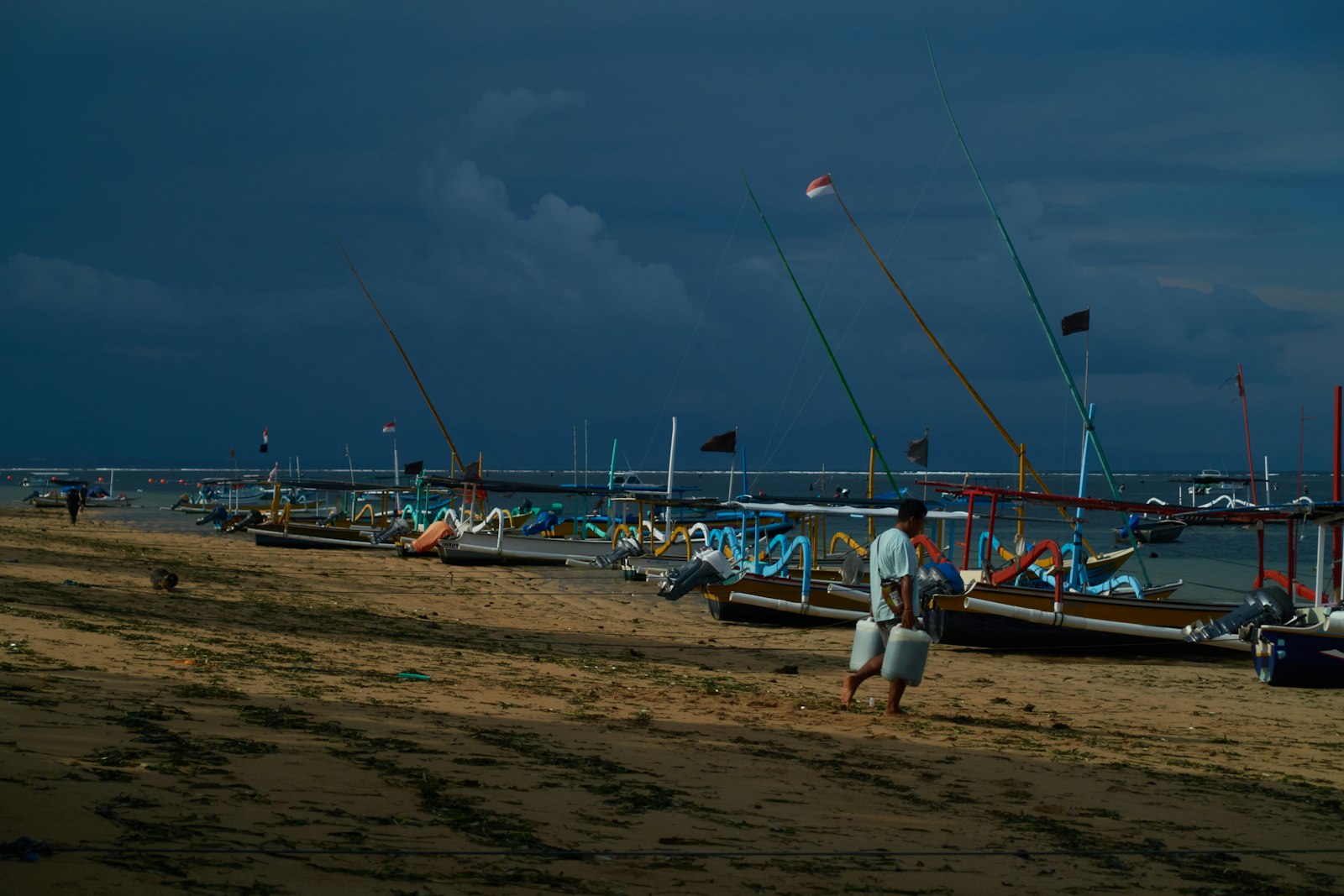 Fishermen with boats on a sandy beach under dark clouds.